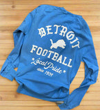 Blue long sleeve shirt on wood floor with large white  print on back. Made in Detroit.