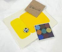 Flat lay of contents from the EK art box, including bold yellow color swatches, geometric print, a pin, and a stamped card.