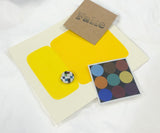 Flat lay of contents from the EK art box, including bold yellow color swatches, geometric print, a pin, and a stamped card.