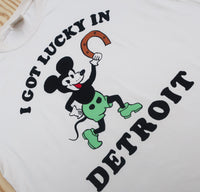 Close-up of cream t-shirt with a retro cartoon holding a horseshoe with black text "I Got Lucky in Detroit."